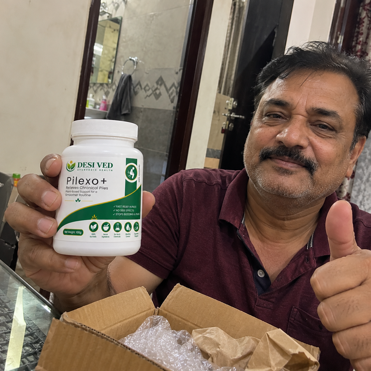 A man holding a white bottle of DESI VED Pilexo+ dietary supplement, giving a thumbs-up while sitting in front of an open cardboard box with bubble wrap inside.